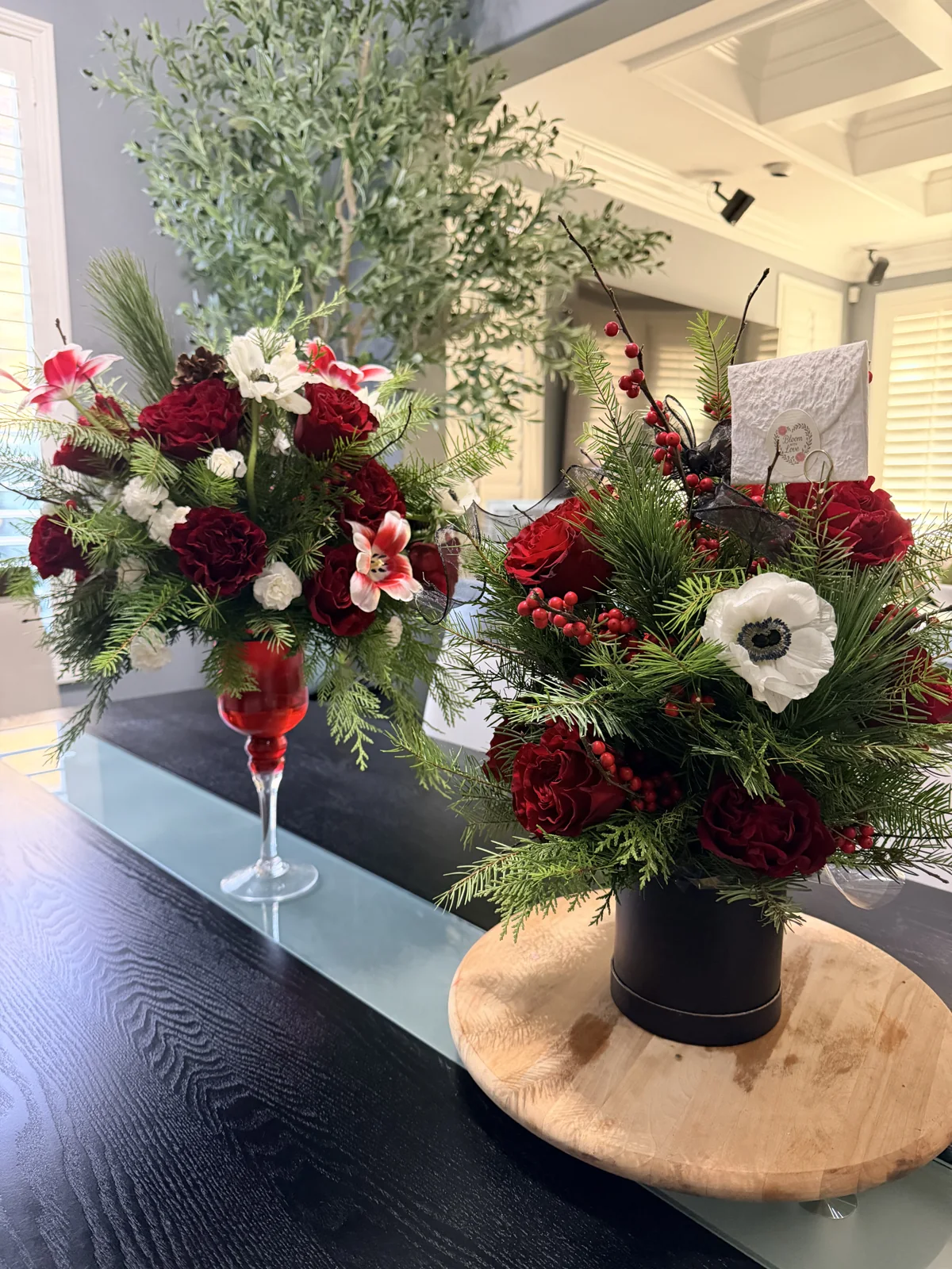 Red roses and white blooms with berries and evergreen foliage in a black flower box beside a red stemmed vessel