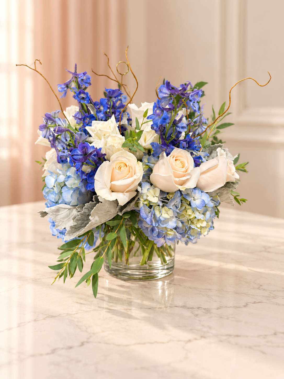 Whispers of Blue arrangement with blue delphinium, soft blue hydrangea, ivory roses, dusty miller and curly willow in a clear glass vase