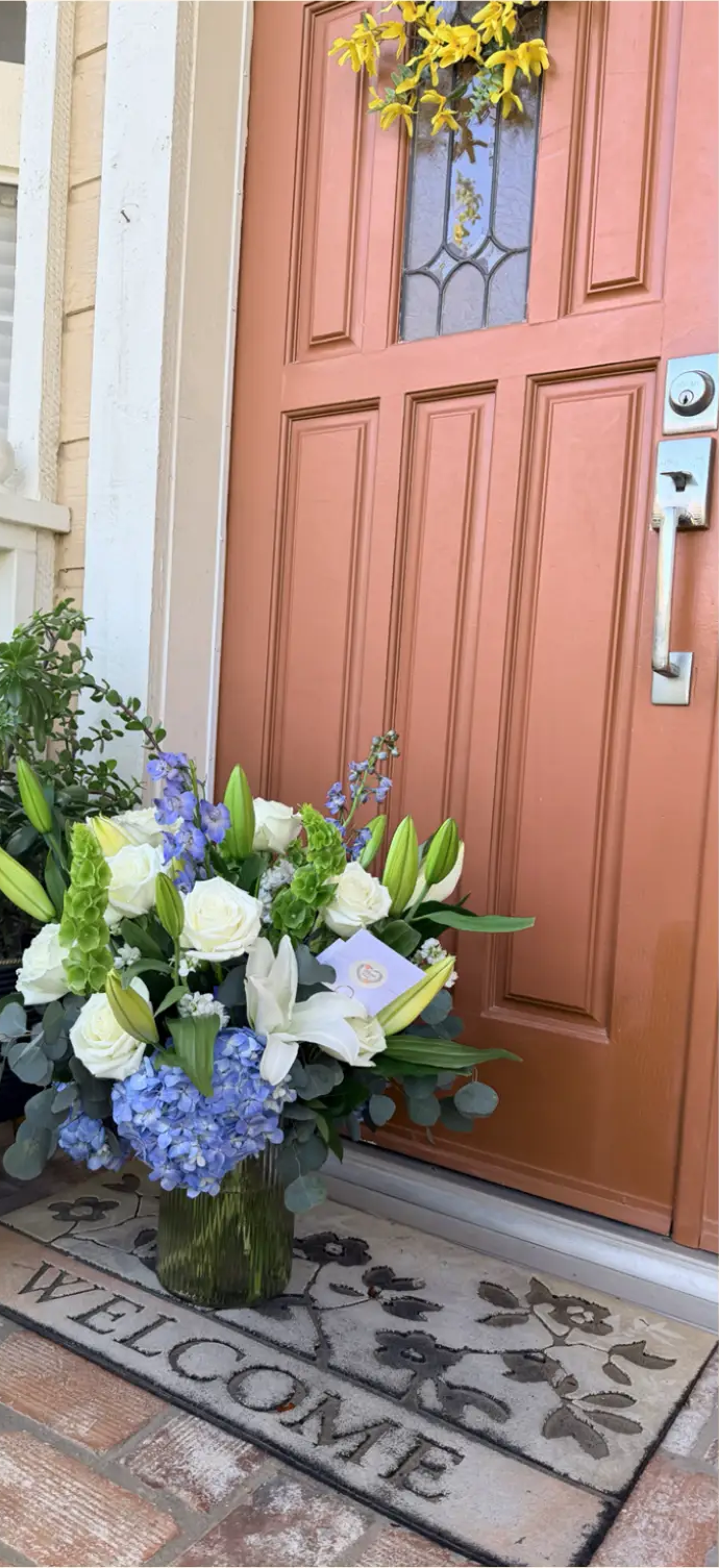Weekly flower subscription delivery of white roses, lilies and blue hydrangeas in a green glass vase on a welcome mat at a front door