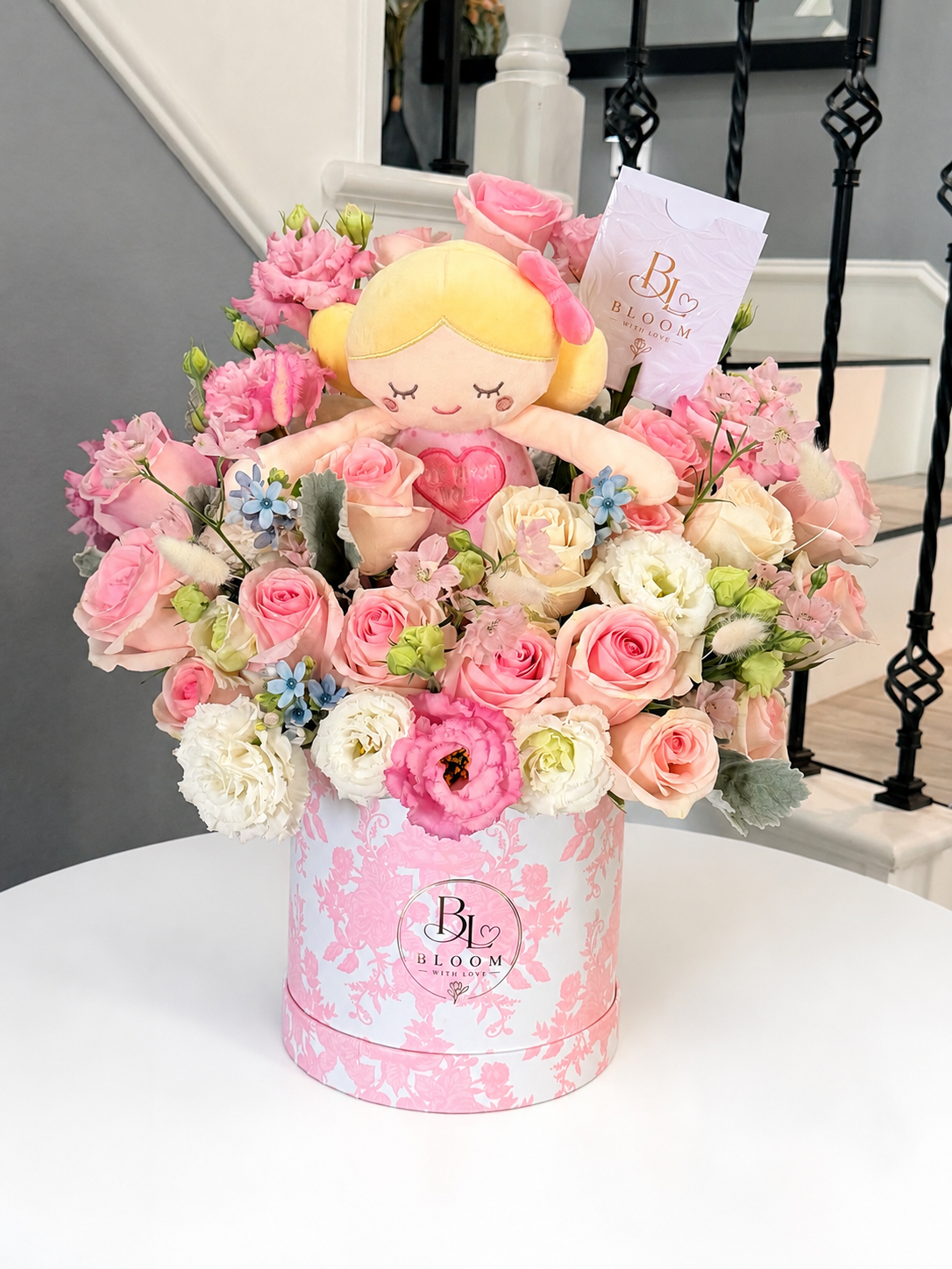 Sweetest Love arrangement of soft pink roses, white lisianthus and blue accents in a pink floral Bloom with Love box with a keepsake doll