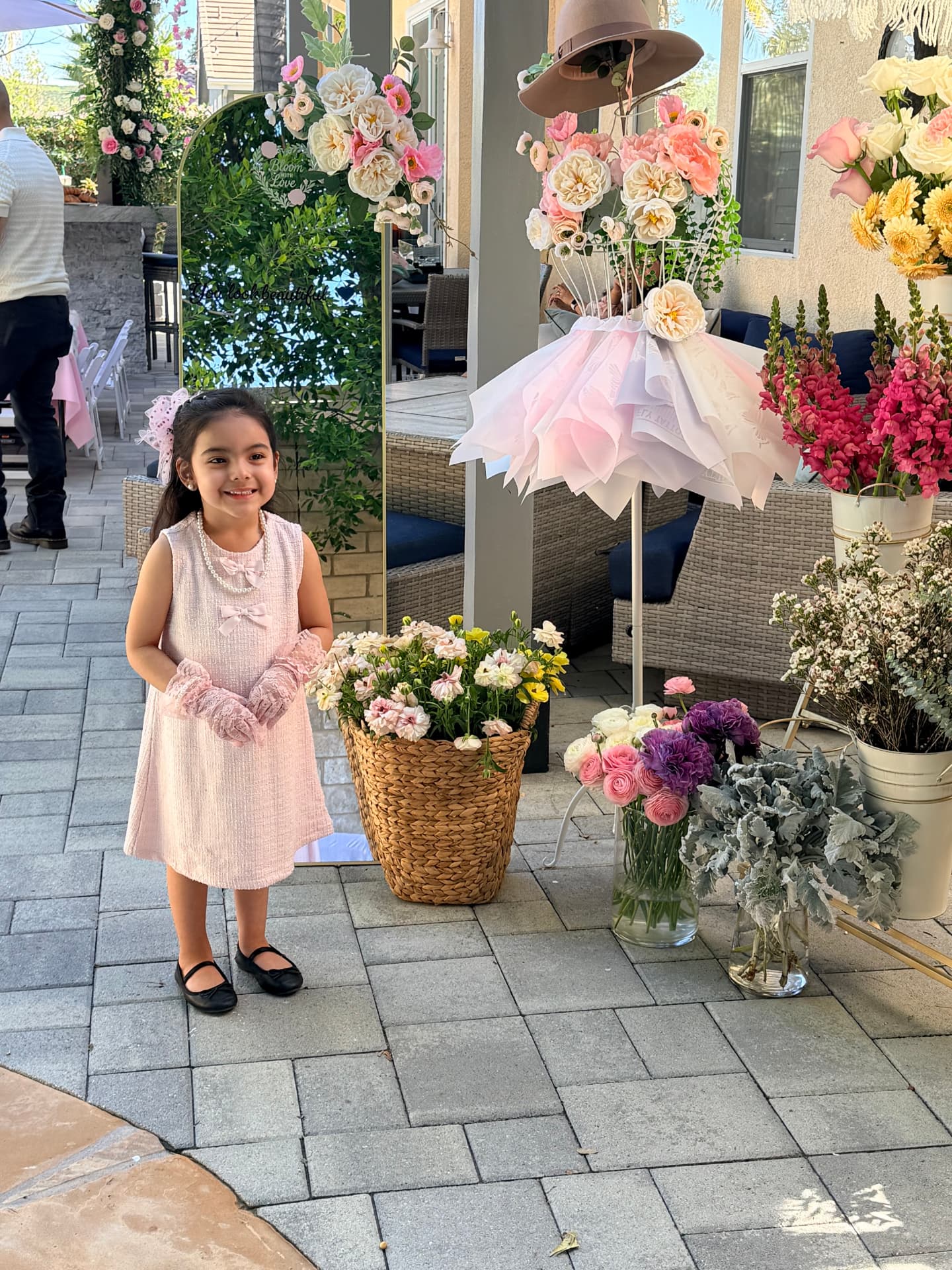 Young guest in a pink dress standing beside the Bloom With Love flower bar and floral mirror at a backyard party