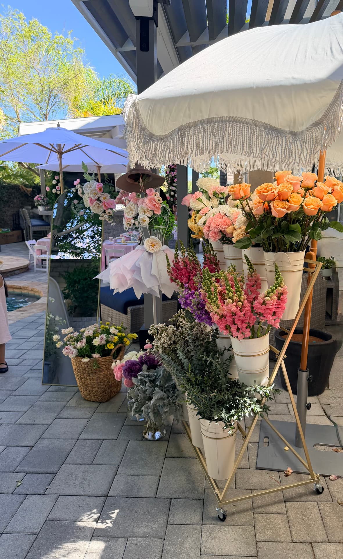 Bloom bar setup poolside with fringed umbrella, gold flower stand and buckets of roses, snapdragons and eucalyptus