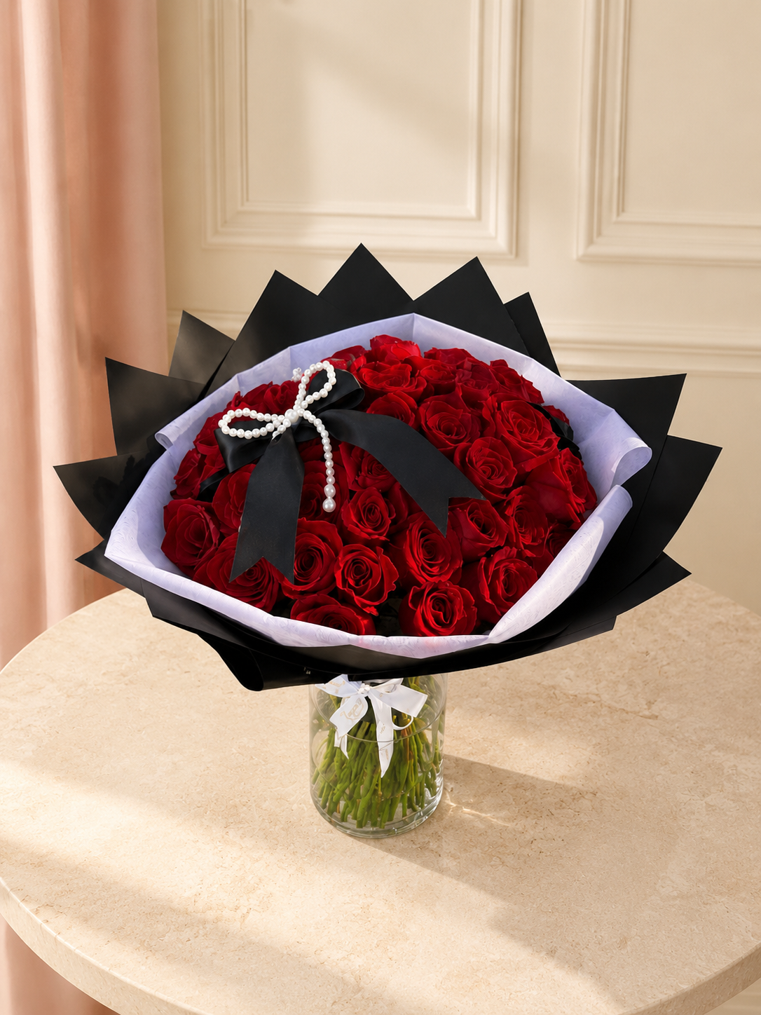 Premium red roses wrapped in dramatic black and soft white paper with a black satin and pearl bow, in a glass vase