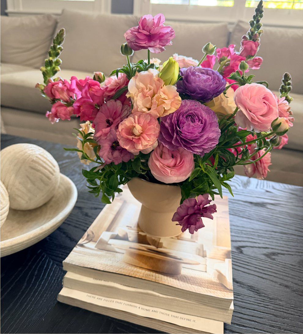 Colorful weekly arrangement of pink, purple and peach ranunculus and snapdragons in a cream pedestal vase styled on a coffee table