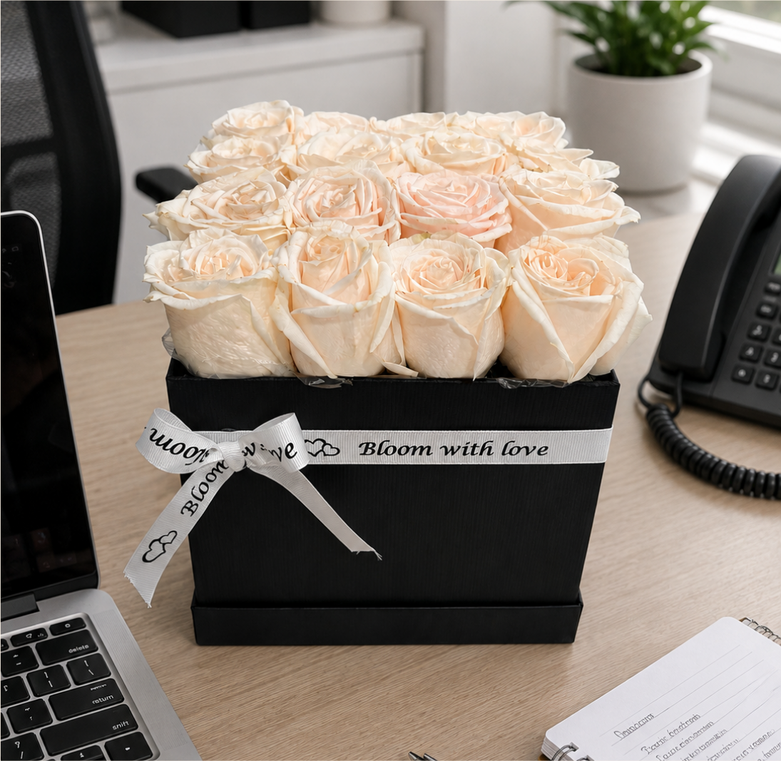 Black keepsake box filled with pale peach roses tied with a Bloom With Love ribbon, styled on an office desk beside a laptop