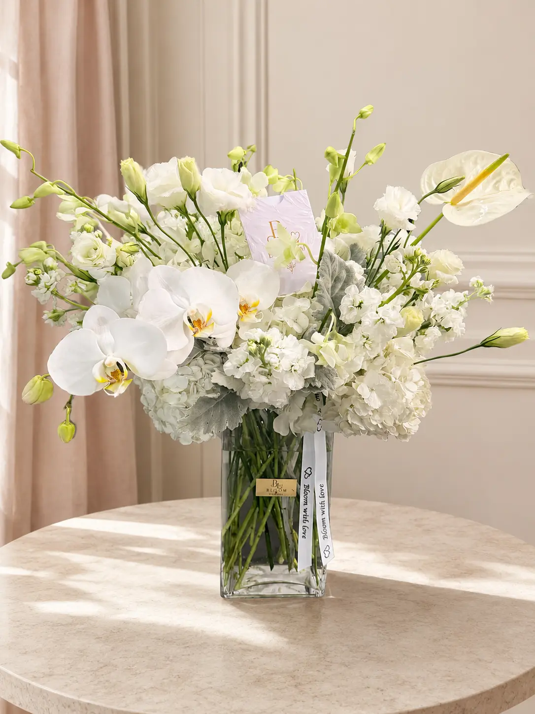 White Elegance tall arrangement with white phalaenopsis orchids, hydrangeas, lisianthus and anthurium in a clear glass vase with Bloom with Love ribbon